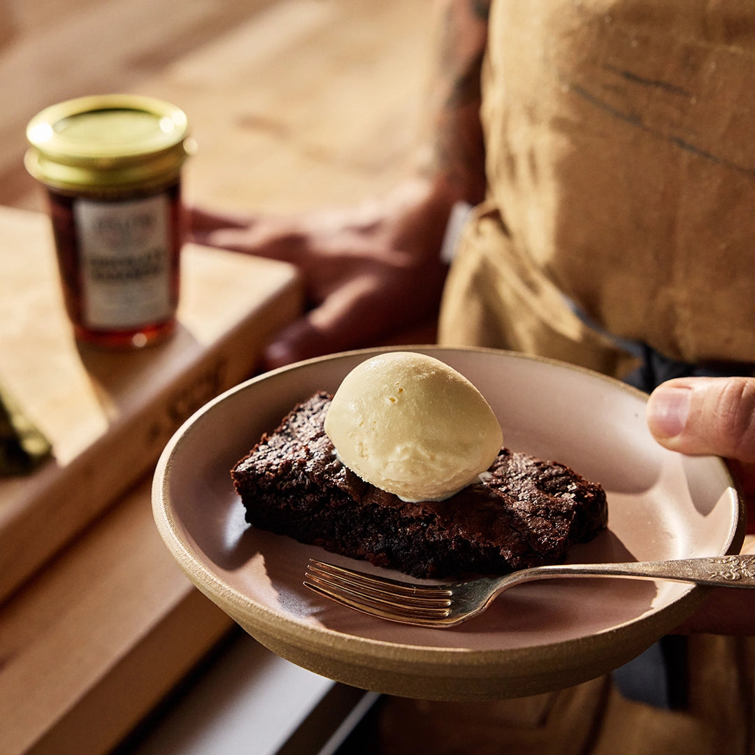 Brownie with ice cream on a plate held by a person, with a jar of pepper jelly in the background.