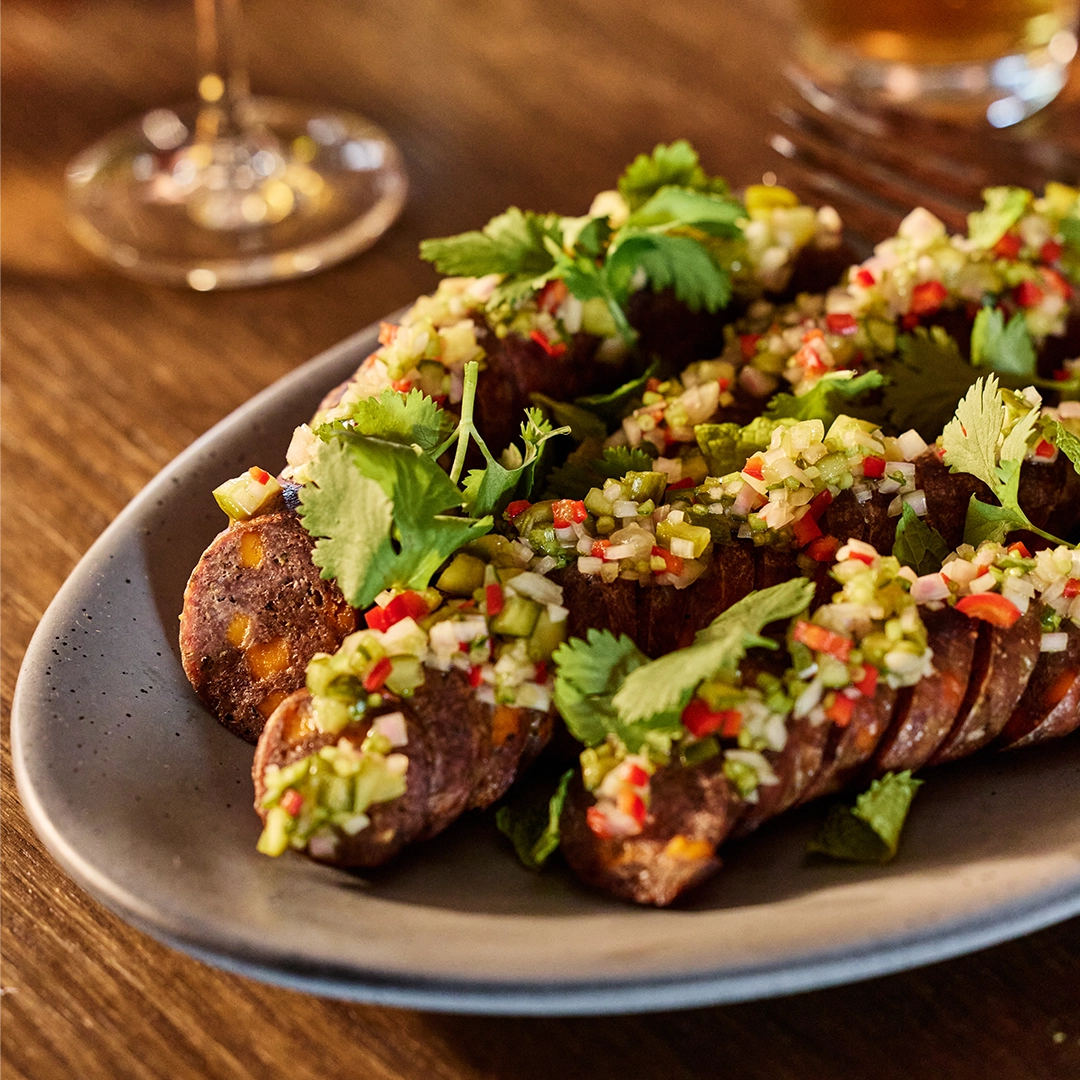 Plated dish of sliced sausage with a green herb and red pepper garnish on a wooden table.