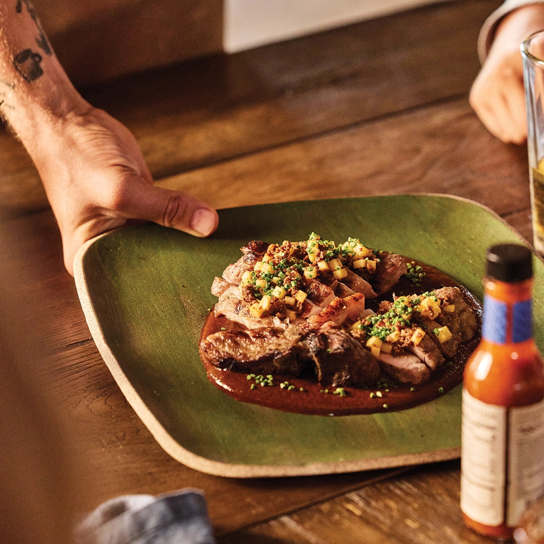 Plated dish of meat with garnish on a wooden table, accompanied by a Stillfire Provisions bottle of hot sauce.