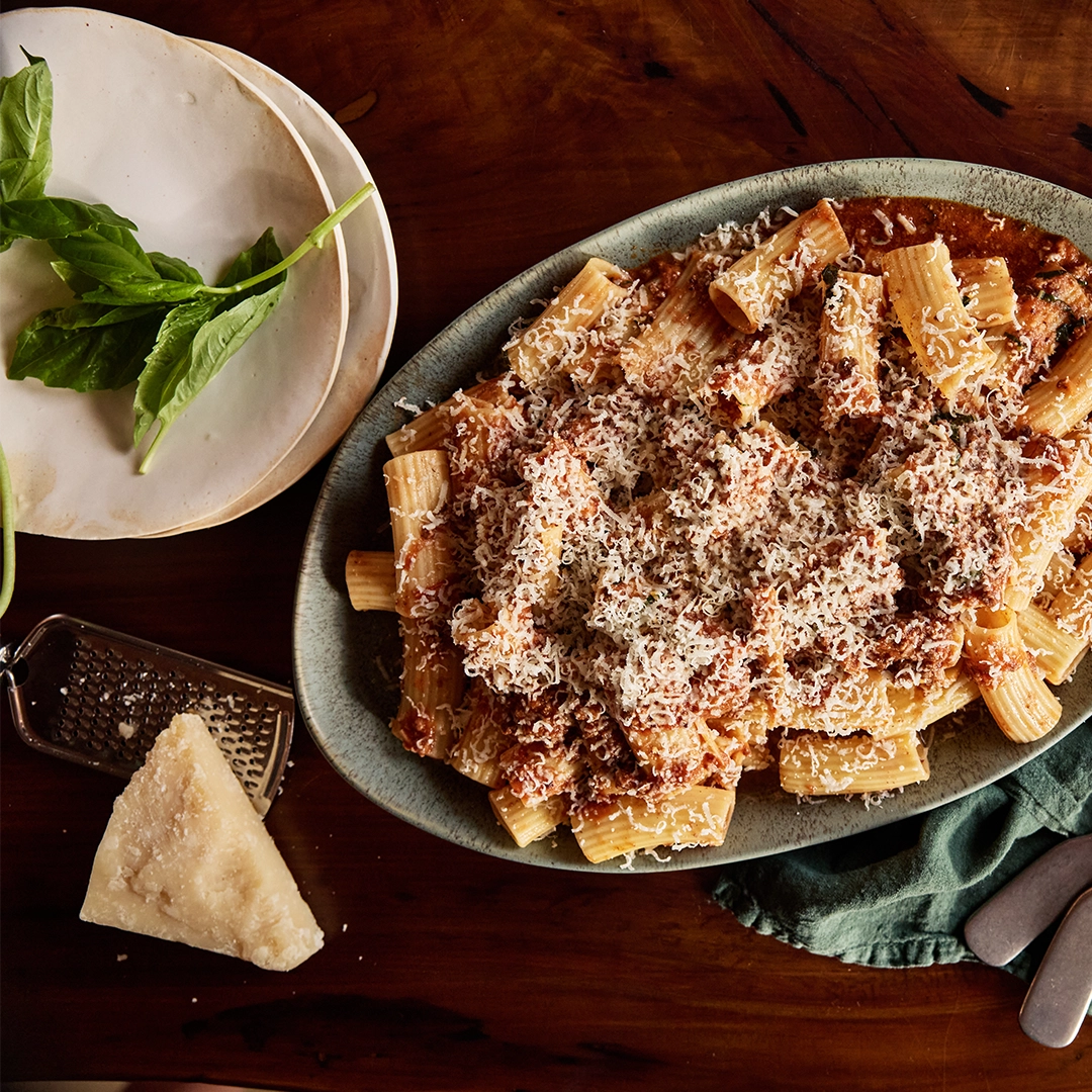Venison pasta dish with tomato sauce and parmesan cheese on a wooden table with a grater and Parmesan cheese.