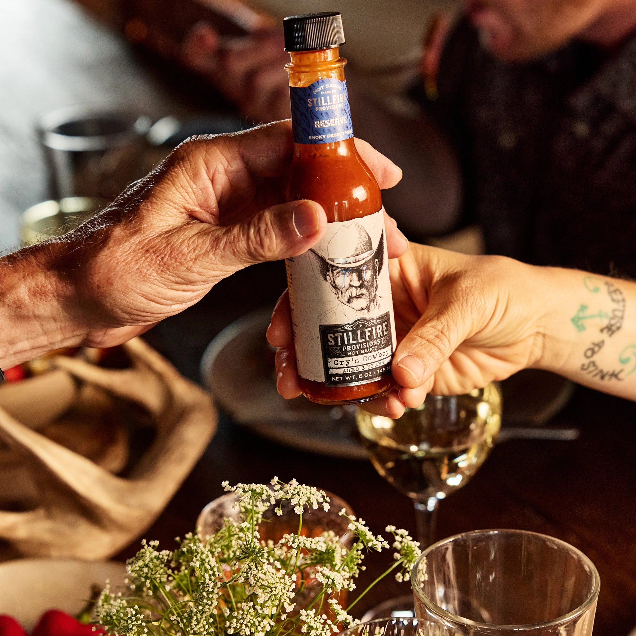 Cry'n Cowboy Hot Sauce being passed by two hands at dinner table.