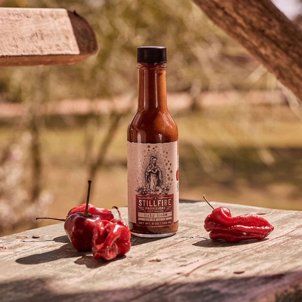 Holy Mole Hot Sauce next to hot peppers on wood table with a country outdoor background.