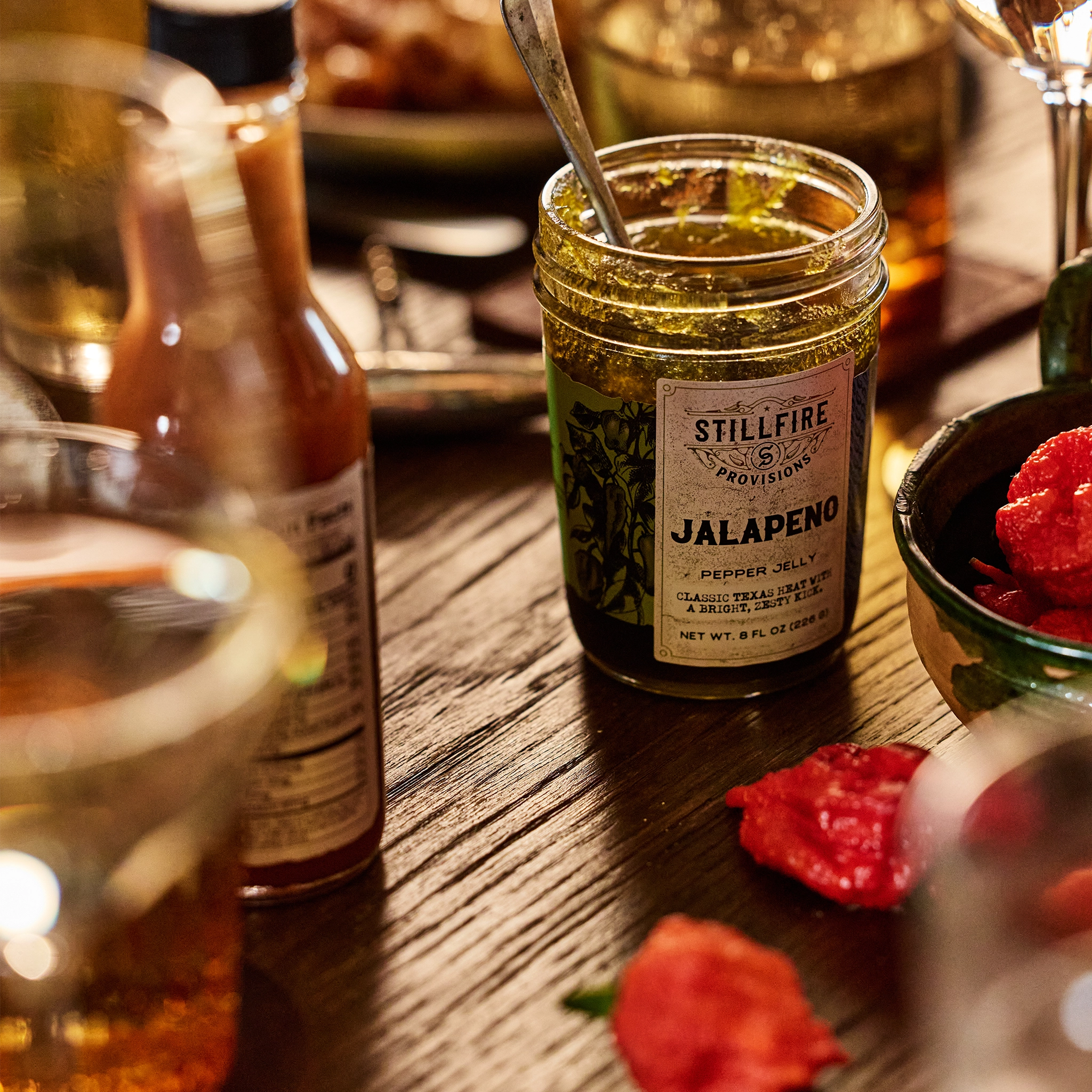 Jalapeno Pepper Jelly with a spoon in the jar on a wood dinner table next to some red peppers.