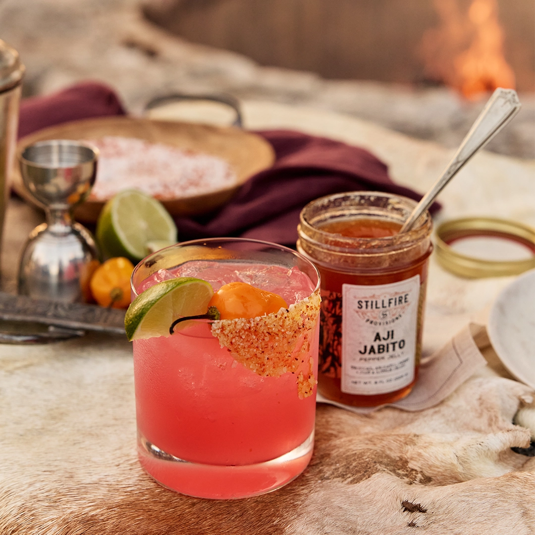 Pink margarita cocktail with lime and orange garnish next to a jar of 'Stillfire Provisions Aji Jabito' on a rustic table.