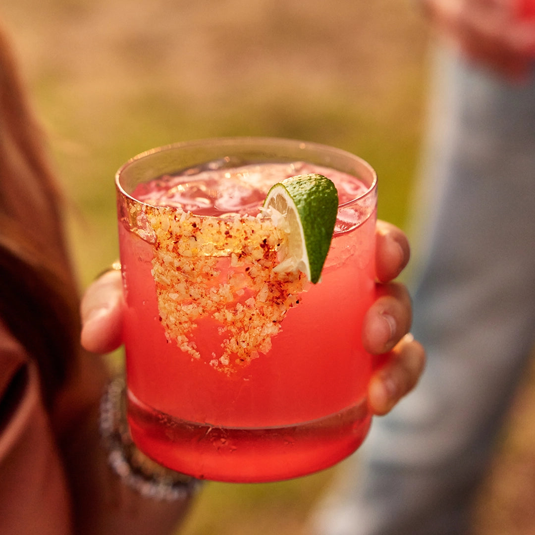 Hand holding a pink cocktail with a lime wedge against a blurred outdoor background