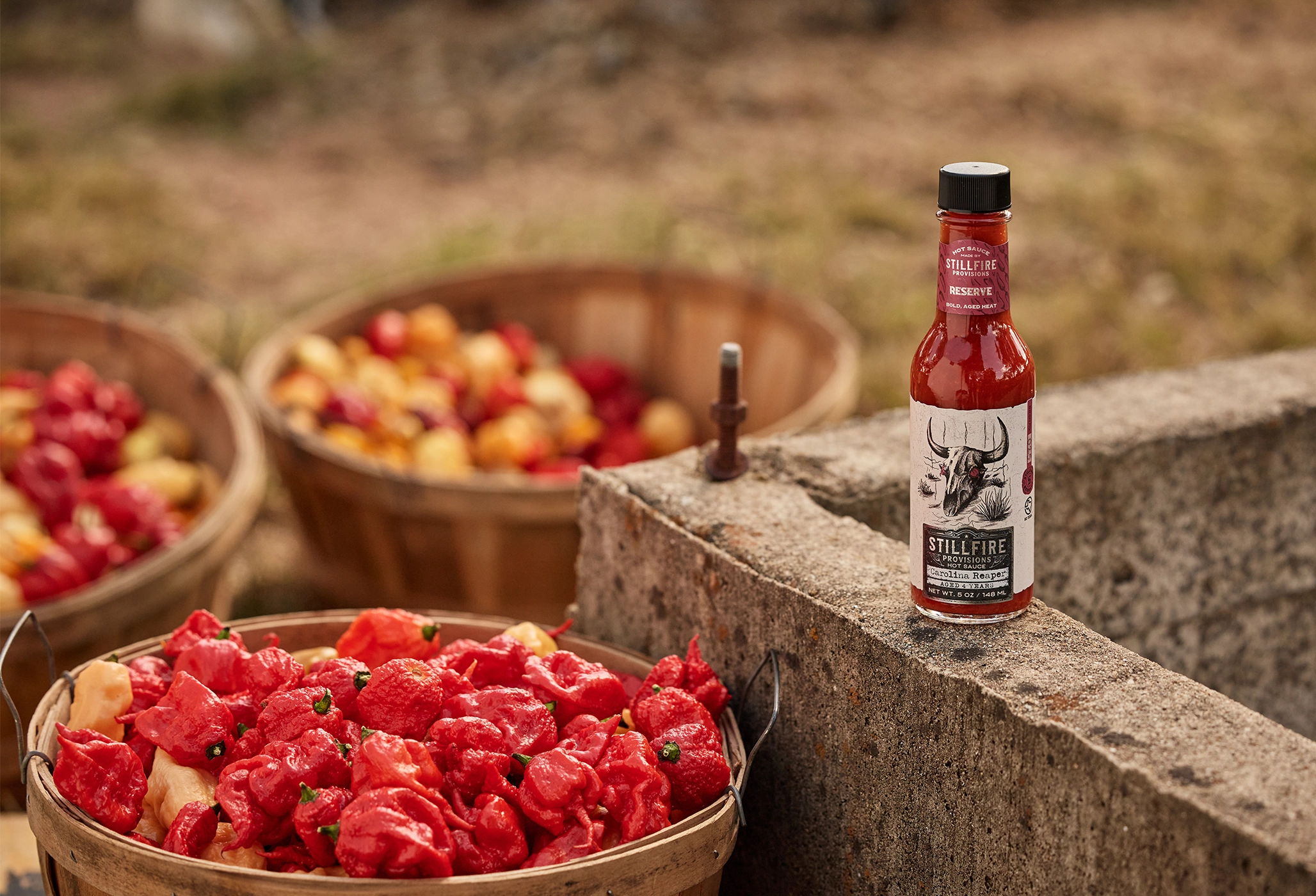 Bottle of hot sauce on a stone ledge with baskets of red peppers and other fruits in the background.