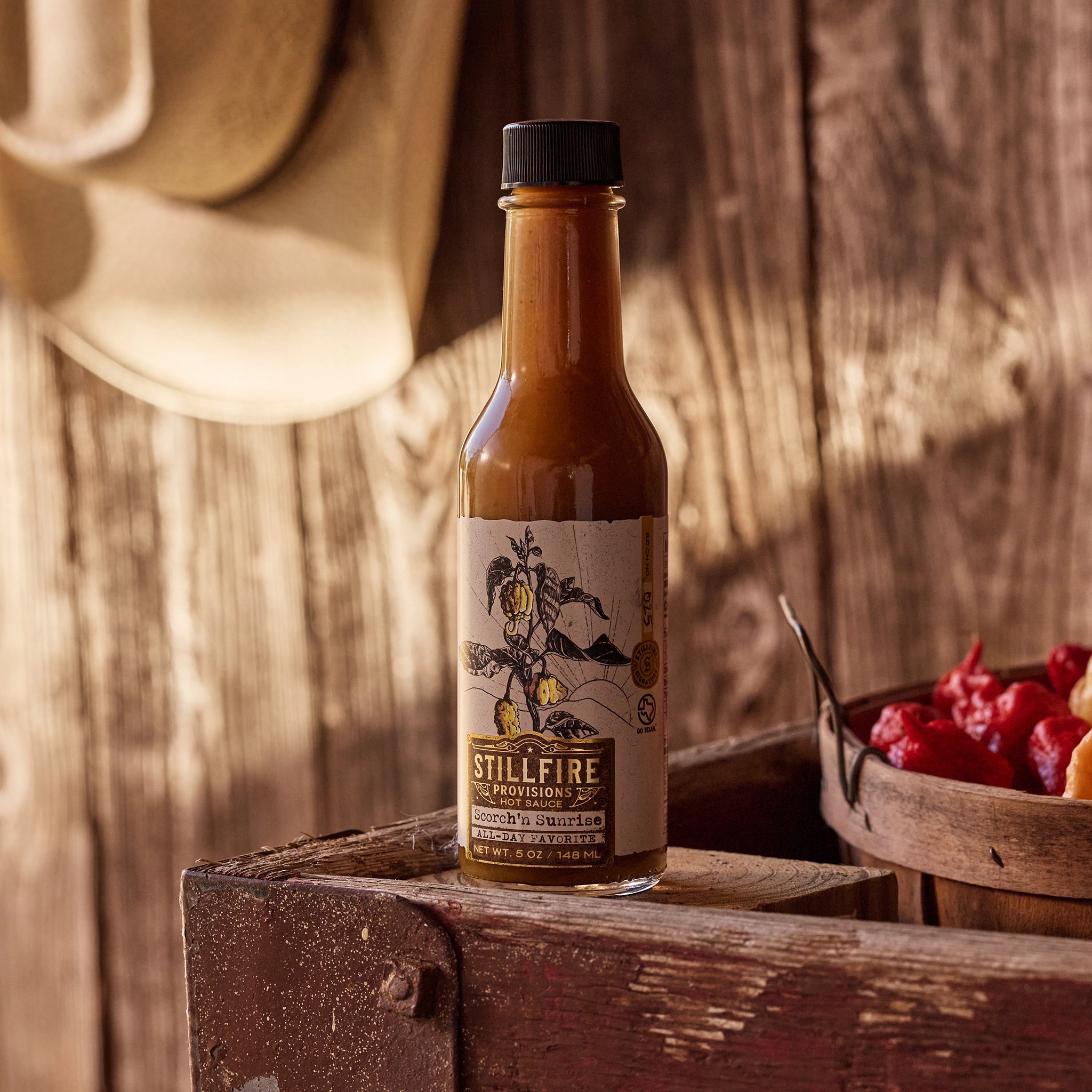 Scorch'n Sunrise Hot Sauce next to a basket of peppers on a wood bench with a cowboy hat in the background.