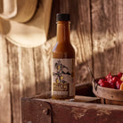 Scorch'n Sunrise Hot Sauce next to a basket of peppers on a wood bench with a cowboy hat in the background.