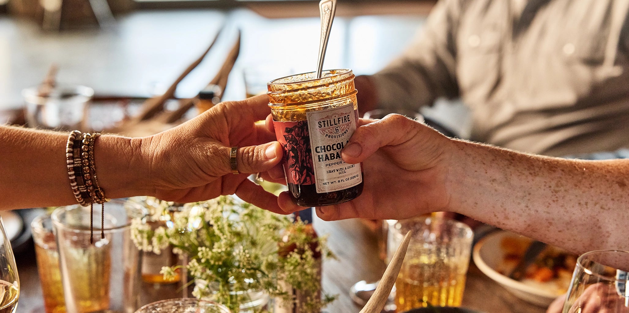 Person holding a jar of chocolate habanero pepper jelly by Stillfire Provisions with another person at a table with drinks and food.