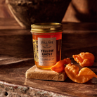 Yellow Ghost Pepper Jelly next to yellow ghost peppers on a wood table with a vase behind it.